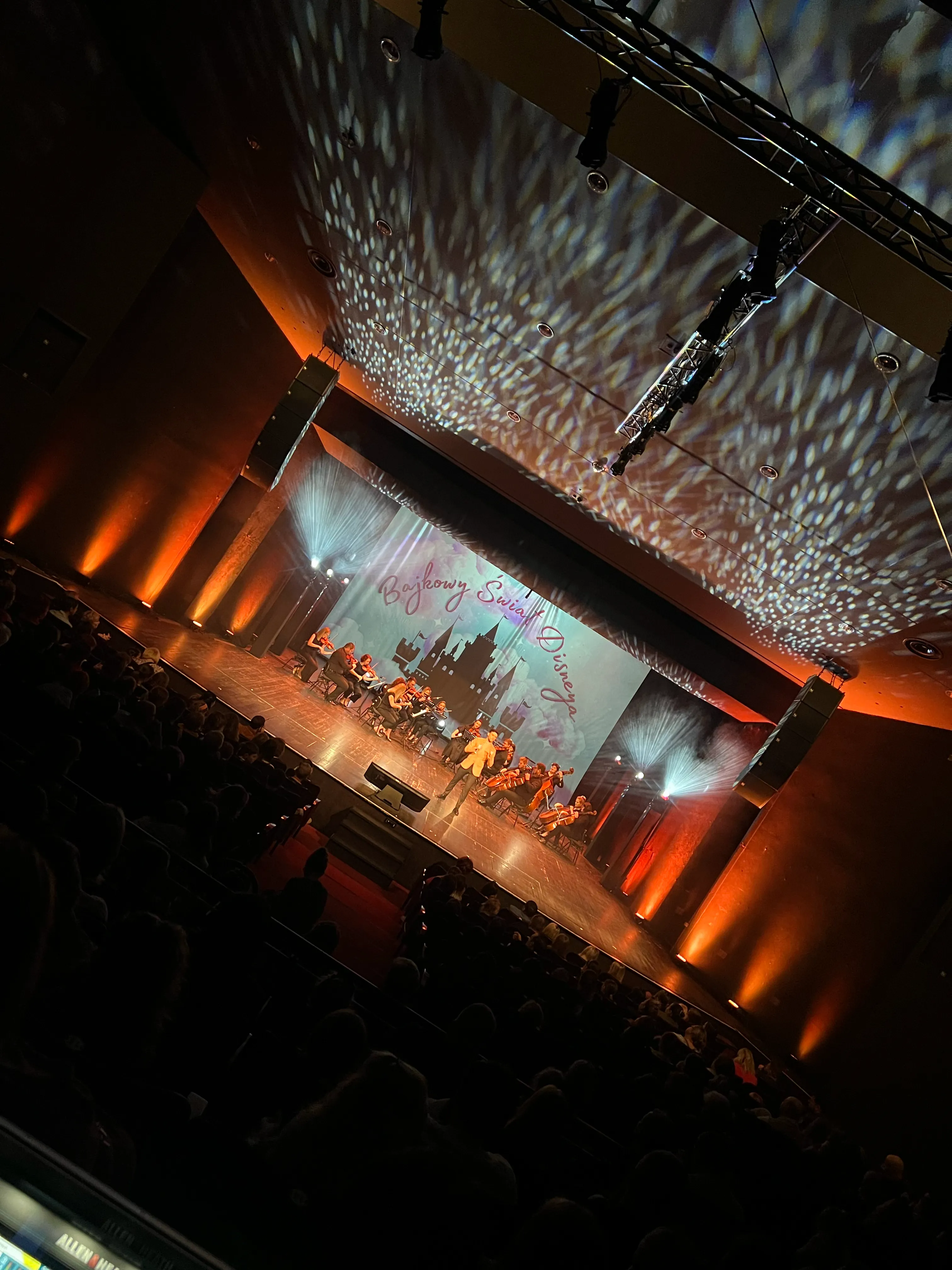 Orchestra and performers on a theatre stage in warm light before a full audience.